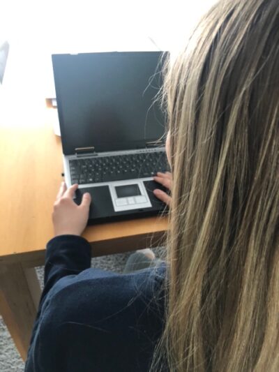 Eine Person mit langen blonden Haaren sitzt an einem Holztisch und arbeitet an einem Laptop. Der Bildschirm des Laptops ist schwarz, und die Person trägt ein dunkelblaues Oberteil. Die Perspektive des Bildes ist von hinten, sodass das Gesicht der Person nicht sichtbar ist.