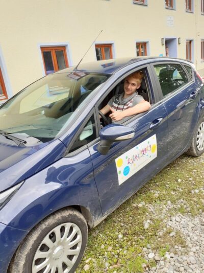 Ein Kind sitzt in einem blauen Auto, das vor einem Gebäude geparkt ist. An der Tür des Autos ist ein buntes Schild angebracht, das offenbar ein Logo oder eine Werbung zeigt. Der Hintergrund zeigt eine beige Wand mit mehreren Fenstern und einem Schild.