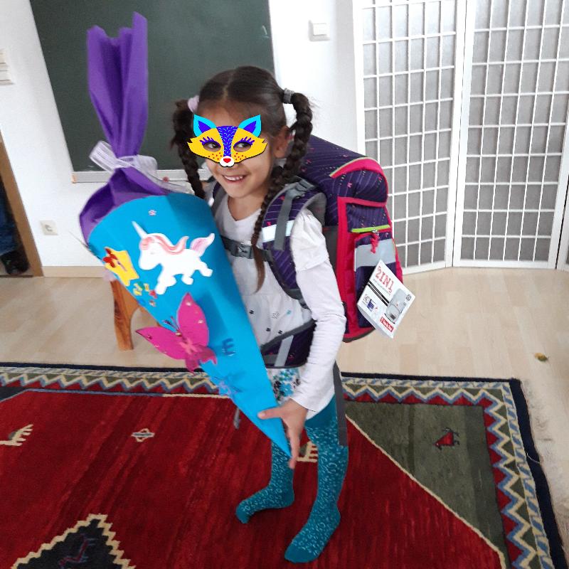 Ein Kind steht in einem Raum und trägt einen großen, bunten Schulranzen auf dem Rücken. In den Händen hält es eine liebevoll gestaltete Schultüte mit Einhorn- und Schmetterlingsmotiven. Der Boden ist mit einem farbenfrohen Teppich bedeckt und im Hintergrund ist eine Tafel und eine weiße Trennwand zu sehen.