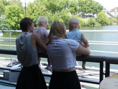 Zwei Erwachsene stehen mit zwei Kleinkindern an einem Geländer und blicken auf einen ruhigen Fluss. Die Umgebung ist grün und bewaldet, und im Hintergrund sind Gebäude zu sehen. Die Szene wirkt friedlich und idyllisch.