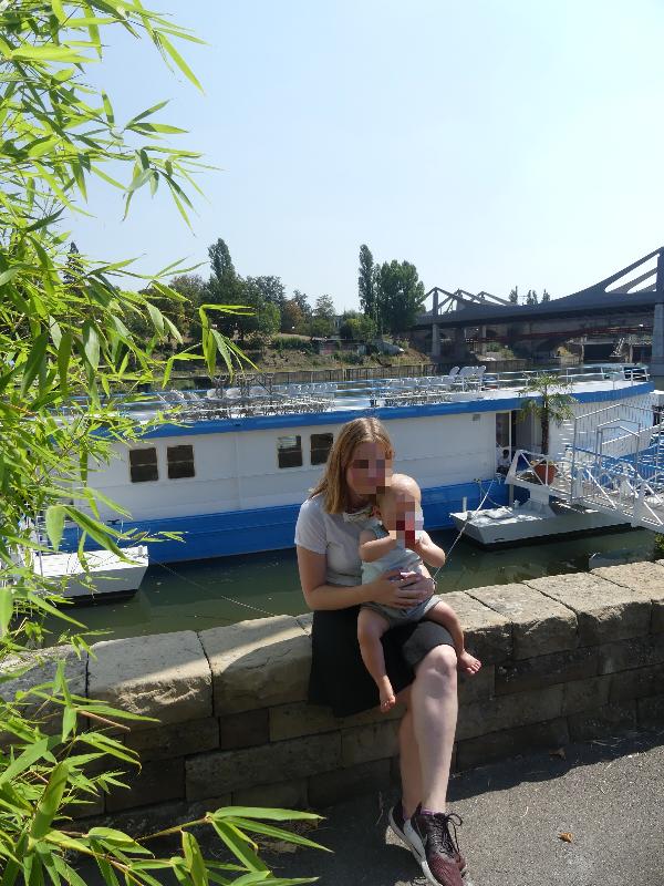 Eine Frau sitzt am Ufer eines Flusses und hält ein Baby im Arm. Im Hintergrund ist ein Boot zu sehen, und sie genießen die ruhige Atmosphäre.