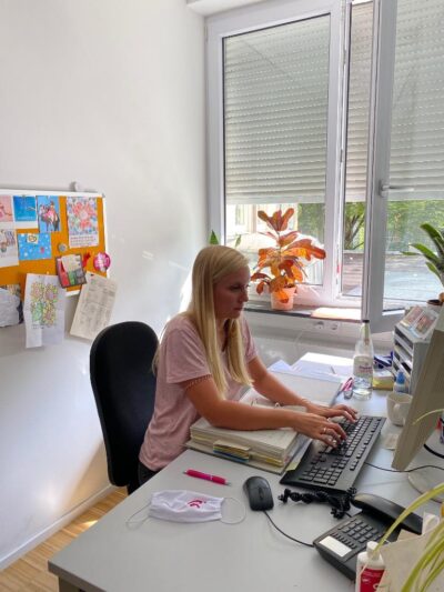 Eine Frau sitzt an einem Schreibtisch und arbeitet am Computer. Der Raum ist hell und freundlich eingerichtet.