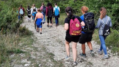 Eine Gruppe von Wanderern geht einen steinigen Pfad entlang, umgeben von grüner Natur. Sie tragen Rucksäcke und sind in Bewegung.