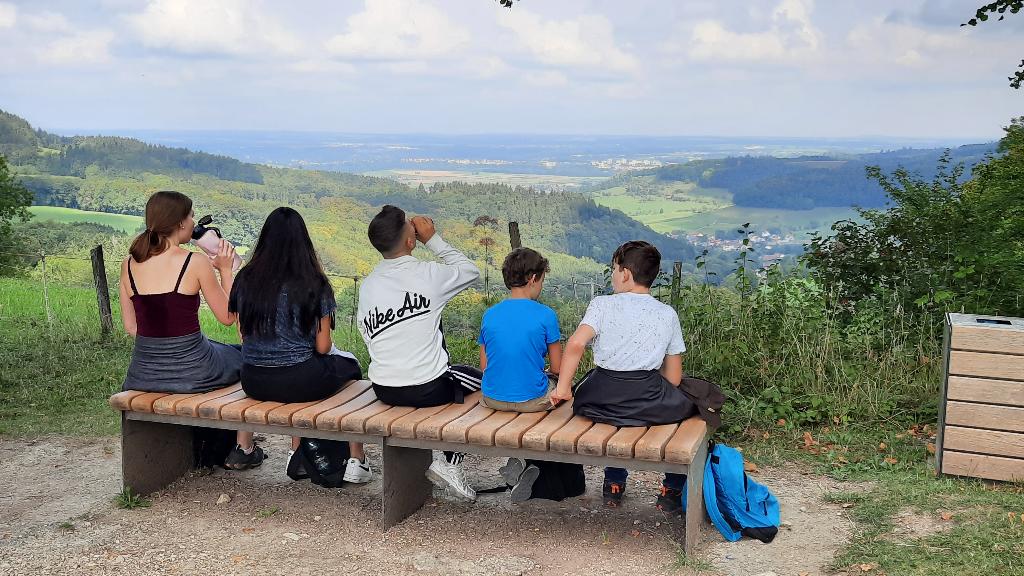 Eine Gruppe von Kindern und Jugendlichen sitzt auf einer Bank und blickt auf eine weite Landschaft. Sie genießen die Aussicht und die gemeinsame Zeit.
