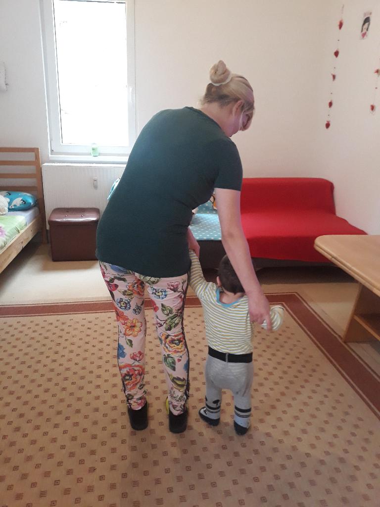 Eine Frau hilft einem kleinen Kind beim Gehen in einem Raum mit Teppichboden. Sie unterstützt das Kind liebevoll und ermutigt es. Sie hält es an den Armen fest, sodass es nicht hinfällt und sich sicher fühlt.