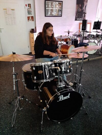 Eine junge Frau spielt Schlagzeug in einem Raum, der wie ein Proberaum aussieht. Sie konzentriert sich auf ihr Instrument und scheint Freude am Spielen zu haben.
