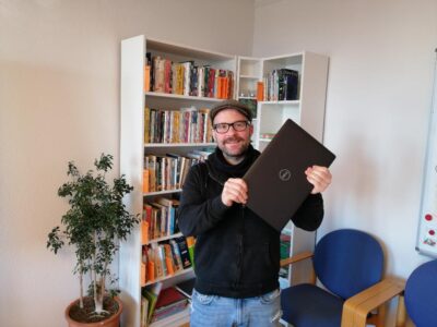 Ein Mann steht in einem Raum und hält einen Laptop in der Hand. Im Hintergrund ist ein Bücherregal mit zahlreichen Büchern zu sehen. Neben dem Regal steht eine grüne Zimmerpflanze in einem Topf. Rechts im Bild sind zwei blaue Stühle und ein Teil einer Pinnwand zu erkennen.