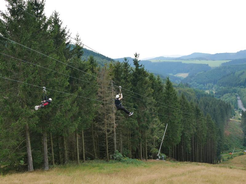 Zwei Personen fahren mit einer Seilrutsche über eine bewaldete Landschaft. Die Szene zeigt dichte grüne Bäume und sanfte Hügel im Hintergrund. Es ist ein aufregender Moment in der Natur, der Abenteuer und Spaß vermittelt.