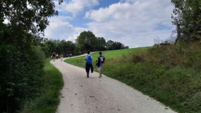 Eine Gruppe von Menschen wandert auf einem geschwungenen, weißen Kiesweg durch eine grüne Landschaft. Zwei Personen im Vordergrund tragen Rucksäcke und gehen nebeneinander. Der Himmel ist teilweise bewölkt, und die Umgebung ist von Bäumen und Grasflächen geprägt.