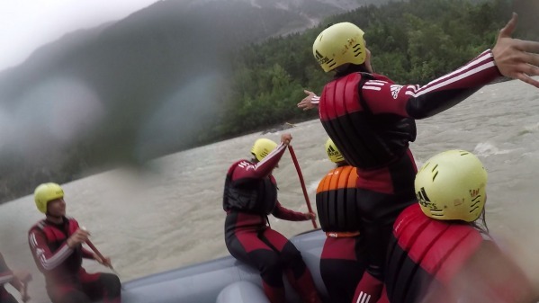Eine Gruppe von Menschen in roten Neoprenanzügen und gelben Helmen ist auf einem Schlauchboot beim Wildwasser-Rafting. Sie navigieren durch raue Gewässer, umgeben von einer üppigen, grünen Waldlandschaft. Die Szene vermittelt ein Gefühl von Abenteuer und Teamarbeit.