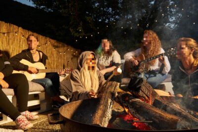 Eine Gruppe von Menschen sitzt um ein Lagerfeuer, während einige von ihnen Gitarre spielen. Die Szene ist von gemütlicher Atmosphäre geprägt, mit Lichterketten im Hintergrund und einem warmen Feuerschein. Die Gruppe scheint einen entspannten Abend im Freien zu genießen.