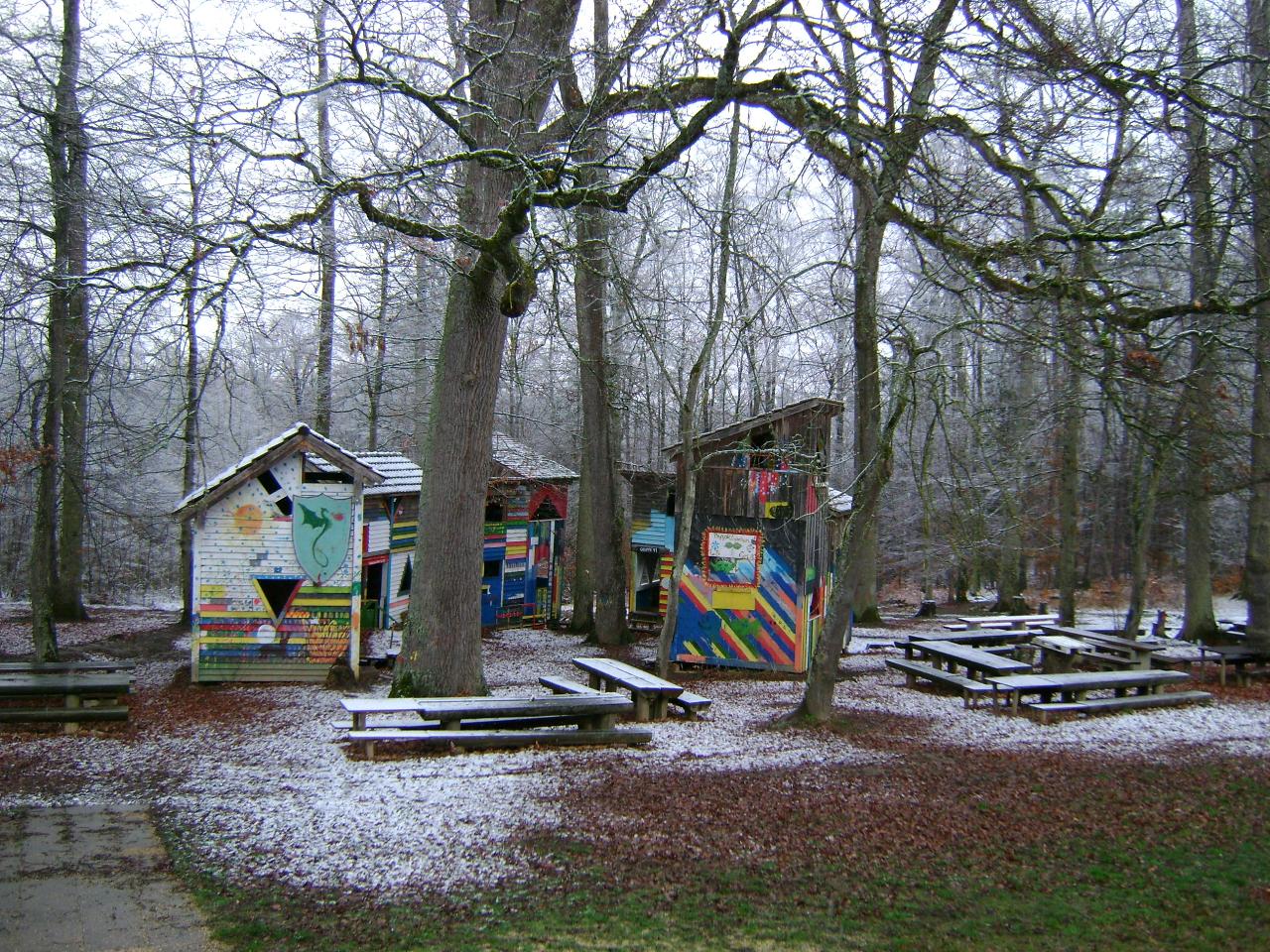 Ein bunt bemaltes Holzhaus steht in einem winterlichen Wald, umgeben von hohen, kahlen Bäumen. Der Boden ist leicht mit Schnee bedeckt und mehrere Holzbänke und Tische sind um das Haus herum angeordnet. Die Szenerie wirkt ruhig und verlassen.