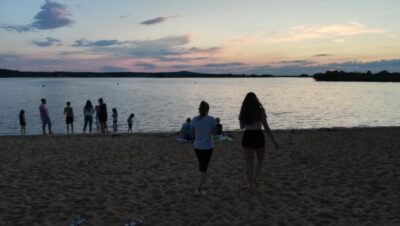 Eine Gruppe von Menschen steht am Ufer eines Sees bei Sonnenuntergang. Der Himmel ist mit sanften Farben von Rosa und Blau gefärbt, während zwei Personen im Vordergrund auf den Strand zulaufen. Die Szene strahlt Ruhe und Gemeinschaft aus.
