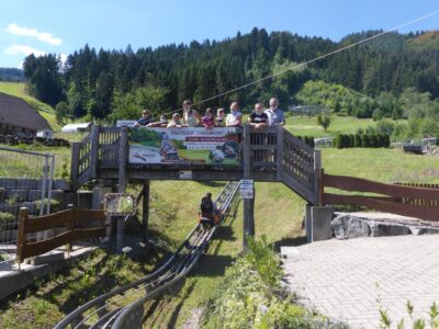 Eine Gruppe von Menschen steht auf einer Holzbrücke und schaut auf eine Sommerrodelbahn hinunter. Im Hintergrund sind grüne Hügel und dichte Wälder zu sehen. Ein Banner auf der Brücke wirbt für ein Kindergeburtstagsangebot.