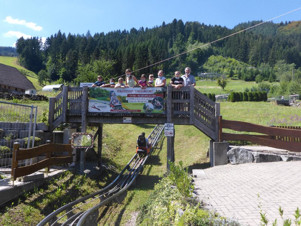Eine Gruppe von Menschen steht auf einer Holzbrücke und schaut auf eine Sommerrodelbahn hinunter. Im Hintergrund sind grüne Hügel und dichte Wälder zu sehen. Ein Banner auf der Brücke wirbt für ein Kindergeburtstagsangebot.
