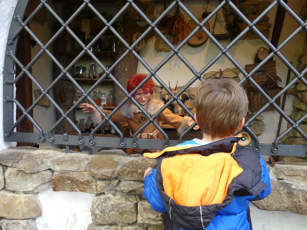 Ein Kind steht vor einem Gitterfenster und schaut in einen Raum, der mit verschiedenen Gegenständen und Dekorationen ausgestattet ist. Im Hintergrund sind Regale mit Gläsern und getrockneten Kräutern zu sehen. Der Raum wirkt wie eine rustikale Werkstatt oder ein Hexenhaus aus einem Märchen.
