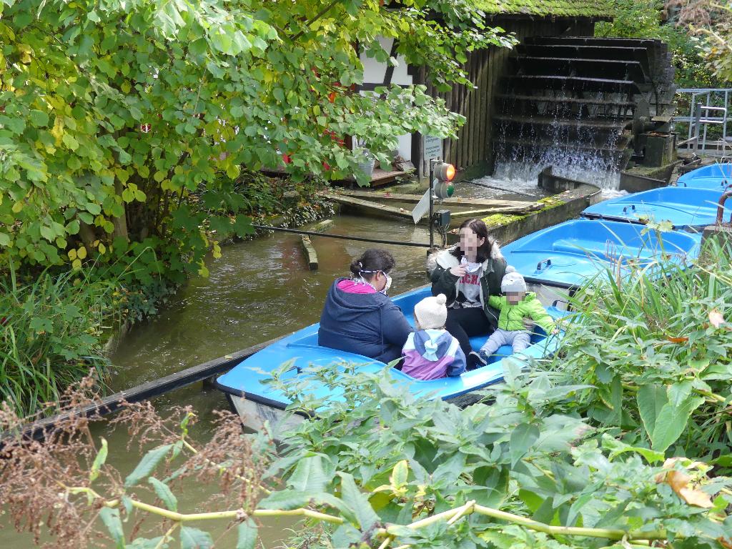 Eine Gruppe von Menschen sitzt in einem blauen Boot auf einem schmalen Fluss, umgeben von üppigem Grün. Im Hintergrund ist ein Wasserrad zu sehen, das Wasser in den Fluss sprudelt. Die Szene wirkt ruhig und friedlich, ideal für eine entspannende Bootsfahrt in der Natur.