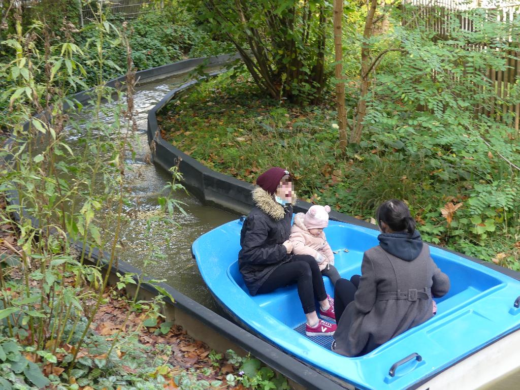 Eine kleine Gruppe von Menschen sitzt in einem blauen Boot und fährt entlang eines schmalen Wasserkanals, der von üppiger grüner Vegetation umgeben ist. Die Szene wirkt ruhig und friedlich, während das Boot gemächlich durch die natürliche Umgebung gleitet. Es ist Herbst, erkennbar an den bunten Blättern, die den Boden bedecken.