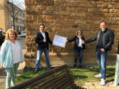 Eine Gruppe von vier Personen steht vor einer Steinmauer im Freien. Zwei der Personen halten gemeinsam ein großes Zertifikat, während die anderen daneben stehen. Im Hintergrund sind ein Gebäude und parkende Autos zu sehen.