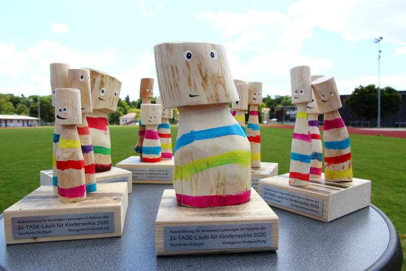Holzfiguren-Trophäen mit bunten Streifen und fröhlichen Gesichtern stehen auf einem Tisch im Freien. Sie sind Auszeichnungen für besondere Leistungen im Rahmen des 24-TAGE-Laufs für Kinderrechte 2020. Im Hintergrund ist eine grüne Wiese und ein Sportplatz zu sehen.