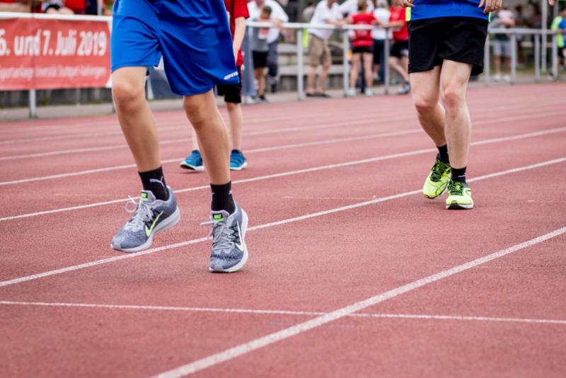 Zwei Personen laufen auf einer roten Laufbahn, wobei nur ihre Beine und Füße sichtbar sind. Beide tragen sportliche Kleidung und Turnschuhe, während im Hintergrund weitere Läufer und Zuschauer zu sehen sind. Ein Banner im Hintergrund deutet auf eine Veranstaltung hin, die im Juli 2019 stattfand.