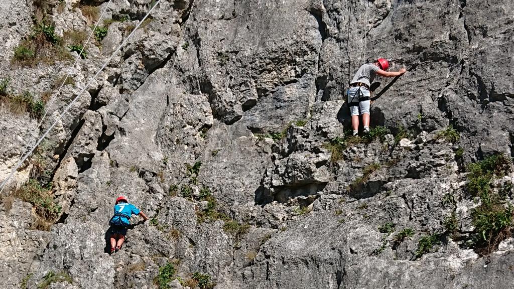 Das Bild zeigt zwei Personen, die eine steile Felswand hinaufklettern. Beide tragen Kletterausrüstung und Helme, während sie sich an den Felsen festhalten. Die Umgebung besteht aus rauem, felsigem Gelände mit einigen grünen Pflanzen, die aus den Spalten der Felswand herauswachsen.