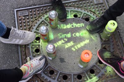 Vier Paar Schuhe umgeben eine bemalte Kanalabdeckung, auf der die Wörter "Mädchen sind cool" und "Girls are strong" in grüner Farbe gesprüht sind. Neben den Schuhen stehen mehrere Spraydosen, die für die Bemalung verwendet wurden. Das Bild vermittelt eine Botschaft der Stärke und Coolness von Mädchen.