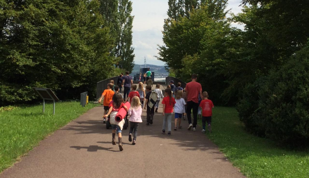 Eine Gruppe von Kindern und Erwachsenen geht einen gepflasterten Weg entlang, umgeben von grünen Bäumen und Gras. Die Kinder tragen bunte Kleidung und einige haben Rucksäcke oder Matten dabei. Im Hintergrund ist eine Aussicht auf eine Stadt mit einem Turm zu sehen.