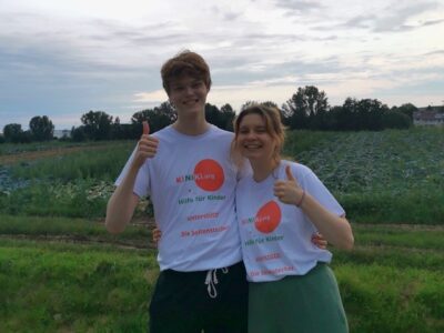 Zwei Personen stehen auf einem Feld und tragen weiße T-Shirts mit dem Logo und Slogan der Organisation KiNiKi.org. Sie zeigen beide den Daumen nach oben und lächeln. Im Hintergrund sind grüne Felder und Bäume unter einem bewölkten Himmel zu sehen.