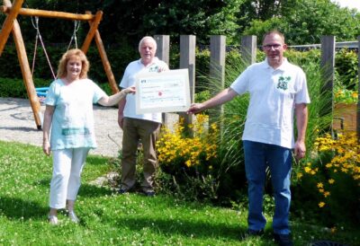 Eine Frau und zwei Männer stehen in einem Garten und halten gemeinsam einen großen Scheck. Im Hintergrund sind eine Schaukel und blühende Pflanzen zu sehen. Die Szene wirkt sommerlich und freundlich.