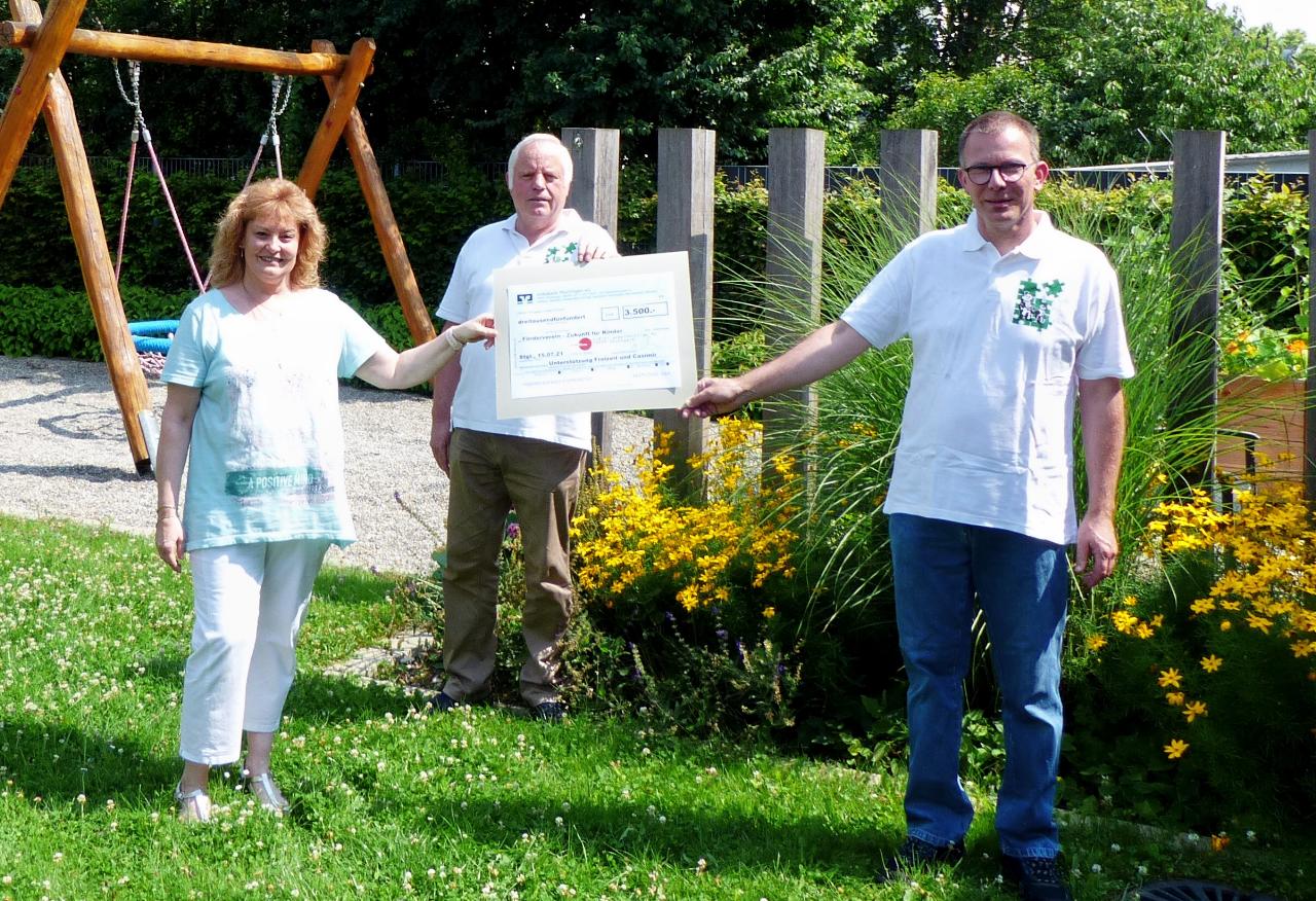 Eine Frau und zwei Männer stehen in einem Garten und halten gemeinsam einen großen Scheck. Im Hintergrund sind eine Schaukel und blühende Pflanzen zu sehen. Die Szene wirkt sommerlich und freundlich.