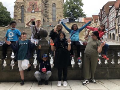 Eine Gruppe von Menschen posiert fröhlich vor einem historischen Gebäude mit einer Uhr und einer großen Treppe im Hintergrund. Sie stehen auf einem steinernen Geländer und machen verschiedene Handzeichen. Im Hintergrund sind traditionelle Fachwerkhäuser zu sehen.