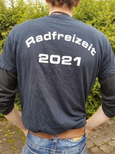 Eine Person steht mit dem Rücken zur Kamera und trägt ein dunkelblaues T-Shirt mit der Aufschrift "Radfreizeit 2021". Die Person hat die Hände in die Hüften gestützt und trägt eine Jeans. Im Hintergrund sind grüne Büsche und ein gepflasterter Boden zu sehen.