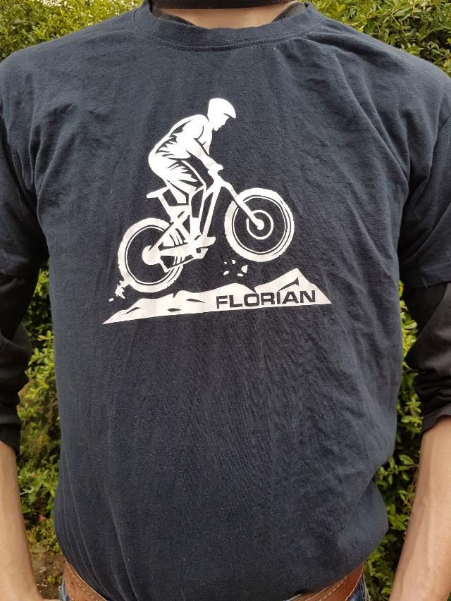Ein Mann trägt ein dunkelblaues Langarmshirt mit einem weißen Aufdruck eines Mountainbikers, der über einen Hügel springt. Unter dem Bild steht der Name "Florian". Im Hintergrund sind grüne Büsche zu sehen.