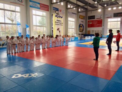 Eine Gruppe von jungen Judokas steht in einer Reihe auf einer roten und blauen Matte in einer Sporthalle. Drei Erwachsene, die vermutlich Trainer oder Betreuer sind, stehen ihnen gegenüber und geben Anweisungen. Im Hintergrund sind große Fenster, Werbebanner und ein Plakat mit der Aufschrift "Judo in Esslingen - Eine Stadt, Ein Team" zu sehen.