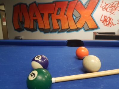 Ein Billardtisch mit blauer Spielfläche und mehreren Billardkugeln, darunter die Nummern 12 und 6, sowie ein Queue im Vordergrund. Im Hintergrund ist eine Wand mit einem großen, bunten Graffiti-Schriftzug "MATRIX" zu sehen. Rechts daneben sind weitere Graffiti-Tags in roter Farbe.