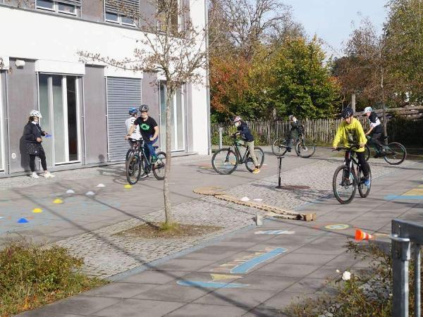 Eine Gruppe von Kindern fährt mit ihren Fahrrädern auf einem gepflasterten Platz vor einem modernen Gebäude. Sie tragen Helme und üben das Fahrradfahren auf einem Parcours mit verschiedenen Hindernissen und farbigen Markierungen. Im Hintergrund sind Bäume und ein Zaun zu sehen, die eine herbstliche Atmosphäre vermitteln.