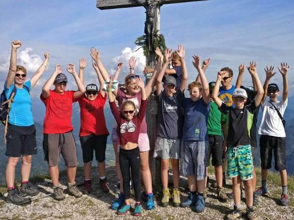 Eine Gruppe von Kindern und Erwachsenen steht auf einem Berggipfel und streckt freudig die Arme in die Luft. Sie sind sportlich gekleidet und tragen Wanderschuhe. Im Hintergrund sind Berge und ein blauer Himmel mit einigen Wolken zu sehen.