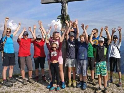 Eine Gruppe von Kindern und Erwachsenen steht auf einem Berggipfel und streckt freudig die Arme in die Luft. Sie sind sportlich gekleidet und tragen Wanderschuhe. Im Hintergrund sind Berge und ein blauer Himmel mit einigen Wolken zu sehen.