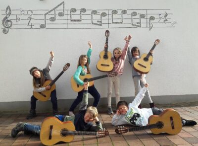 Eine Gruppe von Kindern posiert fröhlich mit Akustikgitarren vor einer Wand, die mit Musiknoten dekoriert ist. Die Kinder zeigen verschiedene kreative Posen und scheinen Spaß zu haben. Der Boden besteht aus Pflastersteinen und die Szene wirkt lebendig und musikalisch.