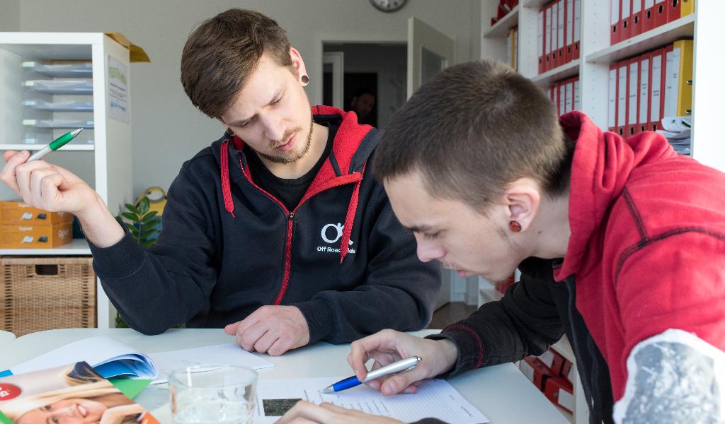 Zwei Personen sitzen an einem Tisch und arbeiten gemeinsam an Dokumenten. Einer der beiden hält einen Stift in der Hand und scheint etwas zu erklären, während der andere konzentriert schreibt. Im Hintergrund sind Regale mit Ordnern und Büromaterialien zu sehen.