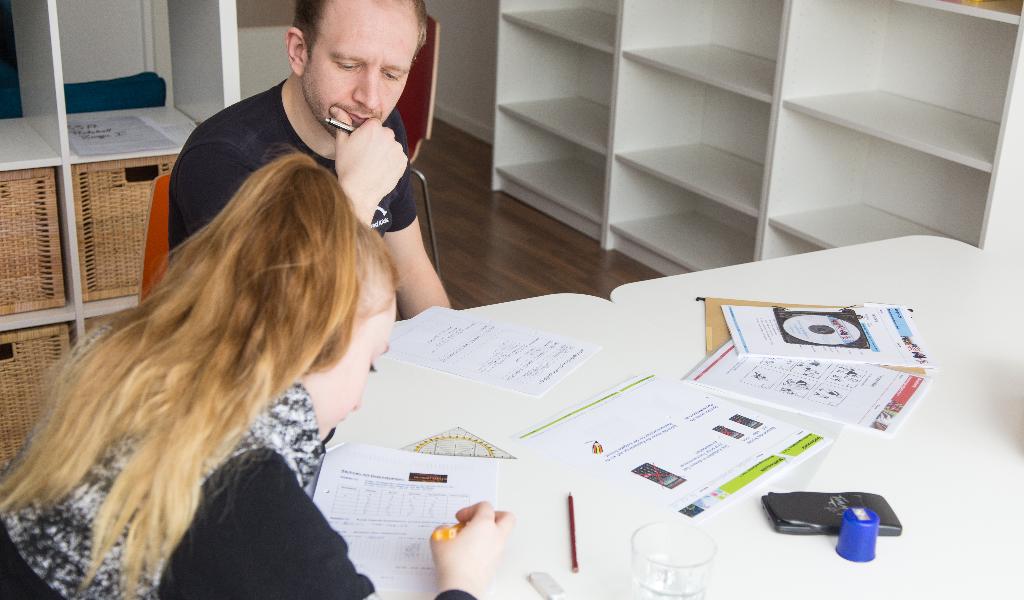 Zwei Personen sitzen an einem weißen Tisch und arbeiten an verschiedenen Dokumenten und Arbeitsblättern. Im Hintergrund sind weiße Regale und Körbe zu sehen. Auf dem Tisch liegen verschiedene Schreibutensilien und ein Handy.