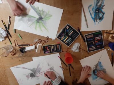 Eine Gruppe von Menschen sitzt an einem Tisch und zeichnet mit Pastellkreiden. Verschiedene bunte Kreidezeichnungen und Zeichenutensilien sind auf dem Tisch verteilt. Hände sind zu sehen, die aktiv an den Kunstwerken arbeiten.