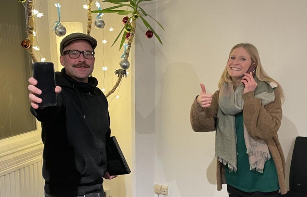 Zwei Personen stehen in einem Raum, der mit weihnachtlichen Dekorationen geschmückt ist. Die Person links hält ein Smartphone und ein Tablet, während die Person rechts einen Daumen hoch zeigt und ein Mobiltelefon ans Ohr hält. Im Hintergrund sind Lichter und Weihnachtskugeln zu sehen, die an einem dekorierten Ast hängen.