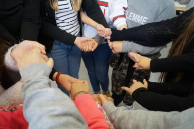 Eine Gruppe von Menschen steht im Kreis und hält sich gegenseitig an den Händen. Die Szene vermittelt ein Gefühl von Gemeinschaft und Zusammenhalt. Die Kleidung der Personen ist bunt und lässig, was auf eine ungezwungene und freundliche Atmosphäre hinweist.