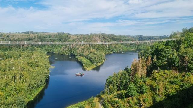 Das Bild zeigt eine malerische Flusslandschaft, umgeben von üppigen grünen Wäldern unter einem blauen Himmel mit einigen Wolken. Über den Fluss spannt sich eine lange Hängebrücke, die sich harmonisch in die natürliche Umgebung einfügt. Ein Boot ist auf dem ruhigen Wasser des Flusses zu sehen, das die friedliche Atmosphäre der Szene unterstreicht.