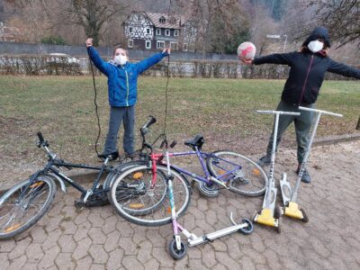 Zwei Kinder stehen neben Fahrrädern und Rollern auf einem gepflasterten Weg. Sie haben ihre Arme in die Luft gestreckt und wirken glücklich und stolz. Im Hintergrund ist eine grüne Wiese und ein Gebäude zu sehen.