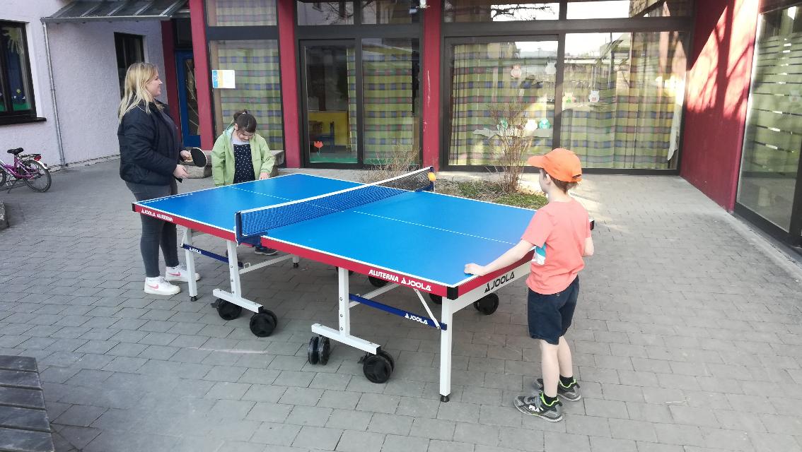Kinder spielen Tischtennis im Freien auf einer blauen JOOLA Tischtennisplatte. Ein Junge in einem orangefarbenen T-Shirt und einer orangefarbenen Mütze steht bereit, während ein Mädchen in einer grünen Jacke den Ball schlägt. Im Hintergrund ist ein Gebäude mit großen Fenstern und bunten Vorhängen zu sehen.