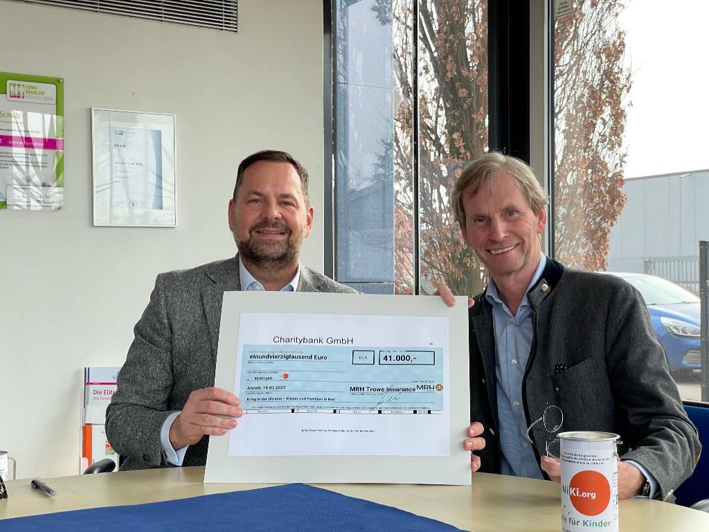 Zwei Personen halten einen großen Scheck über 41.000 Euro, der von der MRH Trowe Insurance an die Charitybank GmbH ausgestellt wurde. Sie sitzen an einem Tisch in einem Büro mit großen Fenstern, durch die Bäume und ein geparktes Auto zu sehen sind. Auf dem Tisch steht ein weißer Becher mit einem orangefarbenen Logo.
