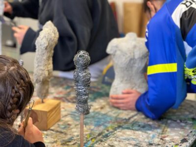 Zwei Personen arbeiten an einer Skulptur aus Stein. Einer von ihnen trägt ein blaues Hemd und hält die Skulptur mit beiden Händen. Auf dem Tisch liegen Werkzeuge und Materialien, die zur Bearbeitung der Skulptur verwendet werden.
